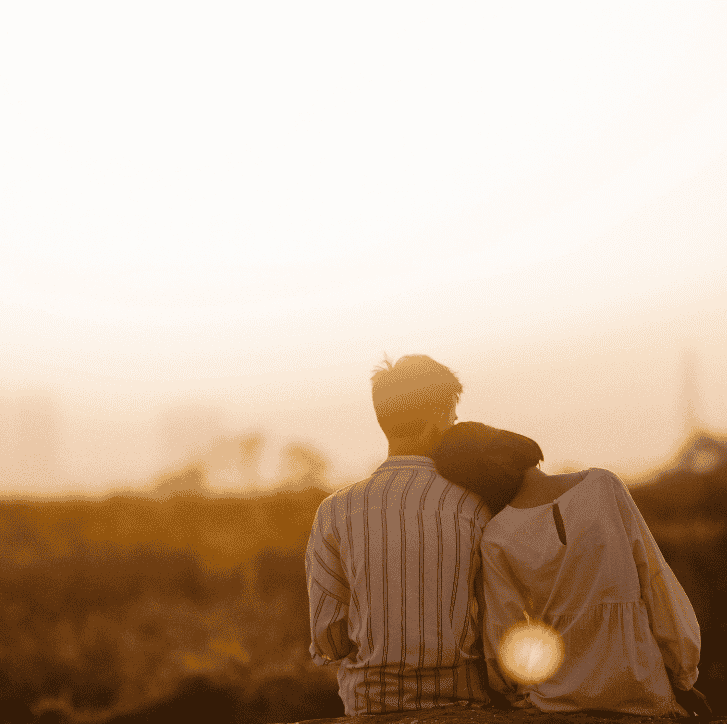 A couple sit closely together, watching a peaceful sunset with their heads resting against each other.